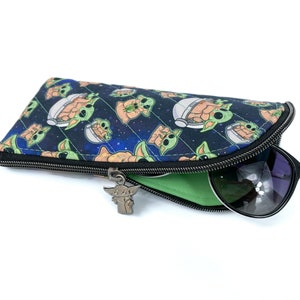 Pu&ograve; includere: Un astuccio per occhiali in tessuto con un motivo spaziale blu scuro che raffigura piccole creature verdi in capsule fluttuanti. L'astuccio presenta una chiusura a zip nera con un ciondolo in metallo a forma di creatura e una fodera verde.