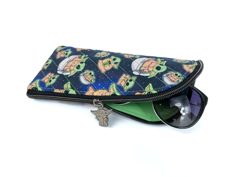 Pu&ograve; includere: Un astuccio per occhiali in tessuto con un motivo spaziale blu scuro che raffigura piccole creature verdi in capsule fluttuanti. L'astuccio presenta una chiusura a zip nera con un ciondolo in metallo a forma di creatura e una fodera verde.