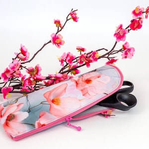 Pu&ograve; includere: Un astuccio rettangolare con cerniera caratterizzato da uno sfondo grigio chiaro con un motivo floreale rosa. L'astuccio ha una cerniera rosa brillante e contiene un paio di forbici nere. Un ramo con fiori rosa &egrave; posizionato dietro.