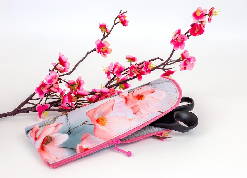 Pu&ograve; includere: Un astuccio rettangolare con cerniera caratterizzato da uno sfondo grigio chiaro con un motivo floreale rosa. L'astuccio ha una cerniera rosa brillante e contiene un paio di forbici nere. Un ramo con fiori rosa &egrave; posizionato dietro.