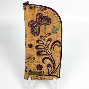 Pu&ograve; includere: Un astuccio alto e curvo in sughero con un motivo a farfalla e fiori viola. L'astuccio presenta una chiusura a zip viola scuro lungo la parte superiore e laterale e una piccola targhetta metallica rettangolare con la scritta handmade.