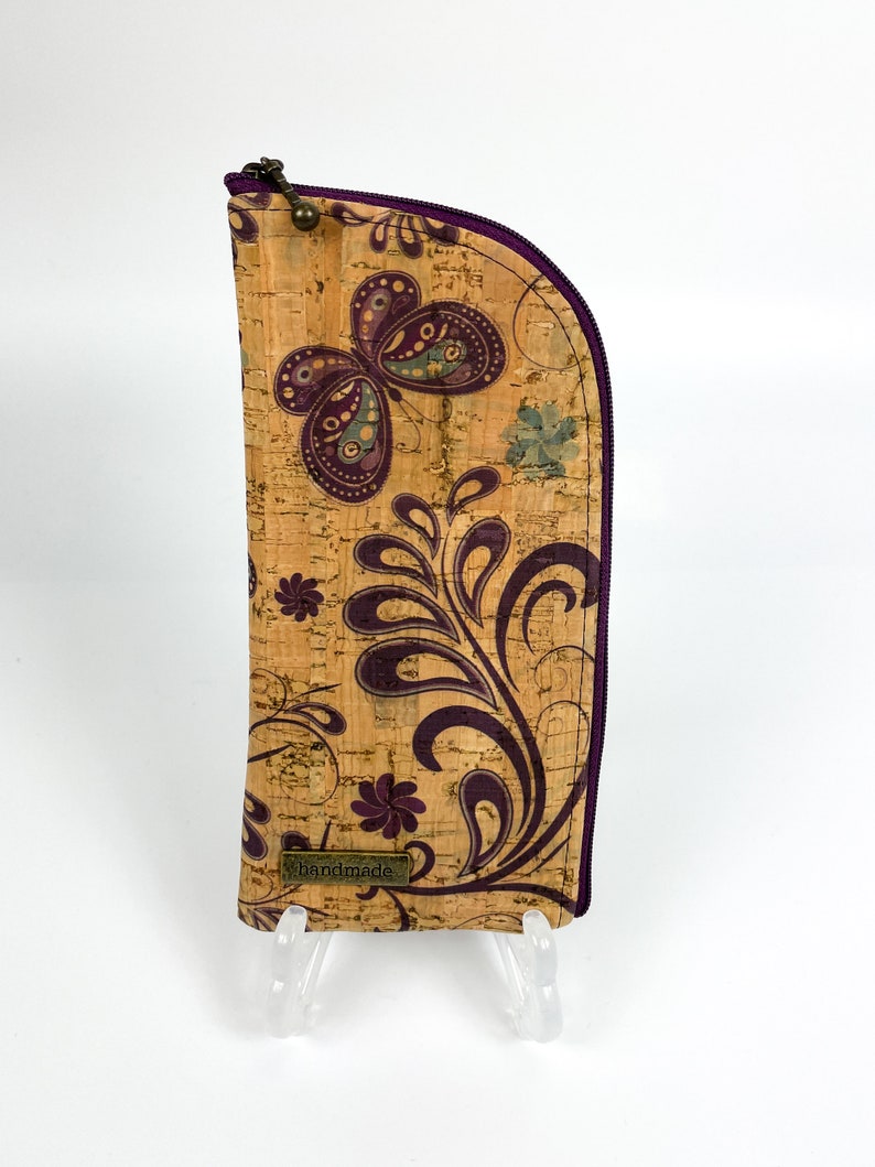 Pu&ograve; includere: Un astuccio alto e curvo in sughero con un motivo a farfalla e fiori viola. L'astuccio presenta una chiusura a zip viola scuro lungo la parte superiore e laterale e una piccola targhetta metallica rettangolare con la scritta handmade.
