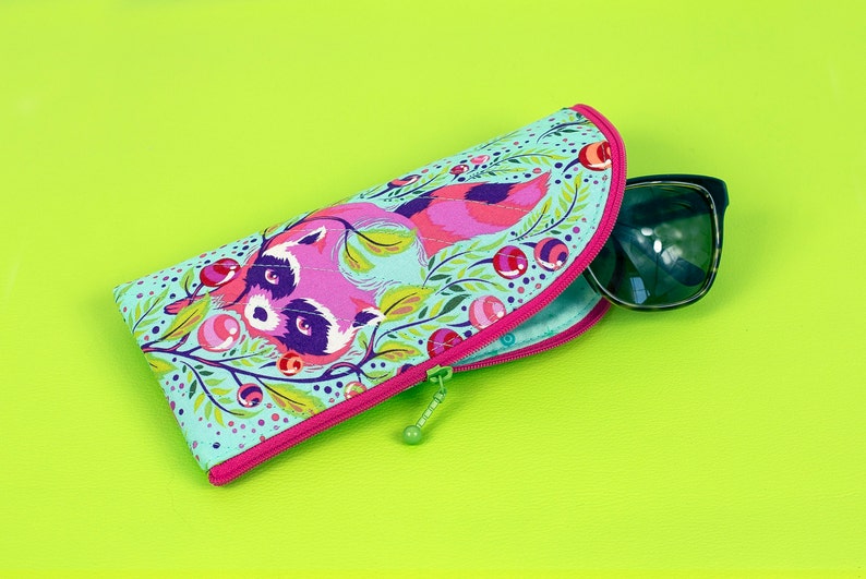 Pu&ograve; includere: Un astuccio per occhiali in tessuto azzurro con una colorata illustrazione di un procione circondato da bacche rosse e fogliame verde. L'astuccio ha una chiusura a zip rosa brillante e contiene un paio di occhiali da sole scuri.