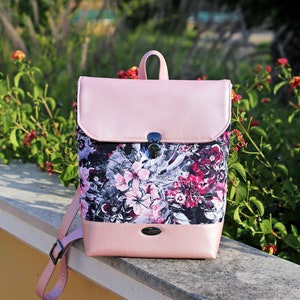May include: A pink floral backpack with a flap closure and a silver buckle. The backpack has a floral pattern with pink, white, and black flowers on a dark background. The backpack is made of faux leather and has a pink strap.