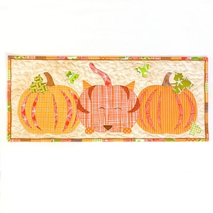 以下が含まれることがあります： 秋をテーマにしたキルトのテーブルランナーには、オレンジ色のカボチャが3つ、そのうちの1つはかわいい猫の顔をしています。カボチャは茶色で縁取られ、格子模様になっています。