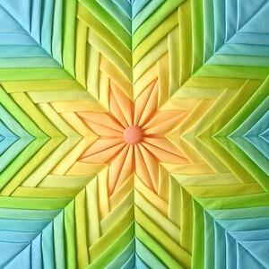 K&ouml;nnte beinhalten: Ein farbenfroher Stoff-Sternenstrahl mit einem zentralen Blumendesign. Der Sternenstrahl besteht aus mehreren Stoffschichten in Blaut&ouml;nen, Gr&uuml;n, Gelb und Orange. Die Blume in der Mitte ist hellorange mit einem rosa Zentrum.