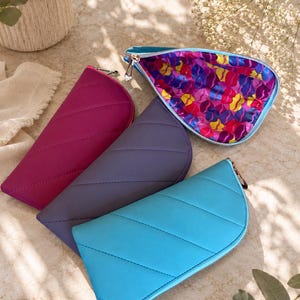Pu&ograve; includere: Quattro pochette semicircolari con cerniera nei colori magenta, grigio scuro e blu brillante. Ogni pochette presenta cuciture trapuntate diagonali e una cerniera color argento. Una &egrave; aperta, rivelando una fodera interna con un motivo astratto.