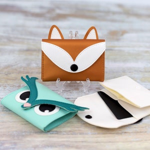 May include: Three small leather wallets with animal designs. A teal blue owl wallet, a brown fox wallet, and a white wallet with a black card holder.