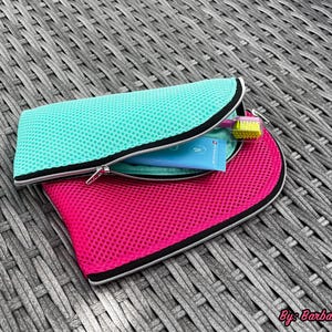 Pu&ograve; includere: Una pochette in rete turchese parzialmente aperta, che rivela un tubetto azzurro e uno spazzolino con setole gialle all'interno, appoggiata sopra una pochette in rete rosa acceso pi&ugrave; grande con bordo nero e cerniera argentata.