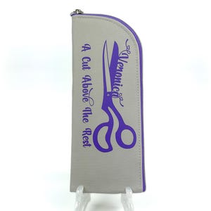 Pu&ograve; includere: Un astuccio grigio a forma di semicerchio con cerniera, caratterizzato da una grafica viola di forbici e dal testo "Veronica" e "A Cut Above The Rest" stampato in viola. L'astuccio presenta un bordo viola lungo la cerniera e un cursore in metallo.