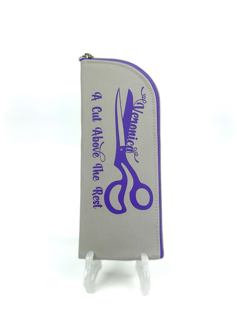 Pu&ograve; includere: Un astuccio grigio a forma di semicerchio con cerniera, caratterizzato da una grafica viola di forbici e dal testo "Veronica" e "A Cut Above The Rest" stampato in viola. L'astuccio presenta un bordo viola lungo la cerniera e un cursore in metallo.