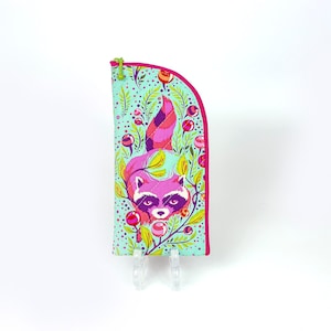 Pu&ograve; includere: Un astuccio in tessuto verticale con bordo superiore arrotondato e cerniera rosa brillante. L'esterno presenta uno sfondo azzurro con un motivo colorato di un procione rosa, fogliame verde e piccole bacche rosse circolari nel design.