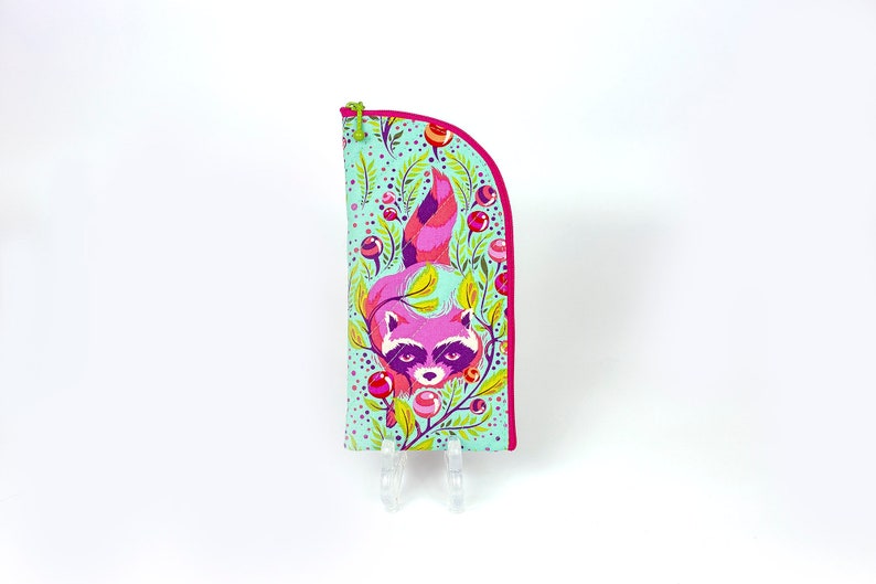 Pu&ograve; includere: Un astuccio in tessuto verticale con bordo superiore arrotondato e cerniera rosa brillante. L'esterno presenta uno sfondo azzurro con un motivo colorato di un procione rosa, fogliame verde e piccole bacche rosse circolari nel design.