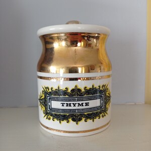 Może przedstawiać: Biały ceramiczny słoik na przyprawy ze złotą pokrywką i czarną i żółtą etykietą z napisem "THYME".