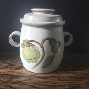 May include: A white ceramic jar with a lid and two handles. The jar is decorated with a green floral design. The lid is a light beige color. The jar is sitting on a wooden surface.