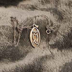 Könnte beinhalten: Silberfarbene Halskette mit einem ovalen Anhänger mit einem keltischen Knotenmuster. Der Anhänger hängt an einer filigranen Kette. Die Halskette liegt auf einer weichen, grauen Oberfläche.