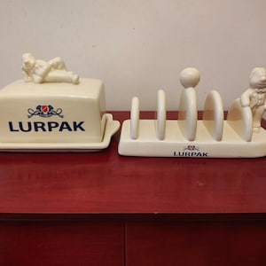 Puede incluir: Juego de mantequera y tostador de cerámica color crema. La mantequera tiene una figura reclinada en la parte superior y la palabra "LURPAK" impresa en la parte delantera. El tostador presenta una figura y la palabra "LURPAK".