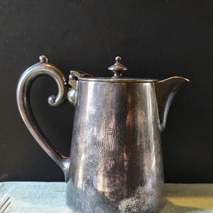 May include: A silver pitcher with a lid and a handle. The pitcher has a monogram on the front.
