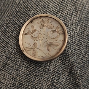 May include: A round, silver-toned pendant with an engraved floral design. The pendant features a detailed pattern of flowers and leaves within a circular frame. The pendant is set against a denim background.