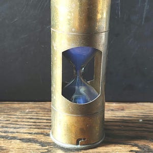 Peut inclure: Un sablier en laiton antique avec un minuteur en verre rempli de sable bleu. Le sablier est enfermé dans un cadre cylindrique en laiton avec une fenêtre de visualisation rectangulaire. Le laiton a un aspect usé et vieilli.