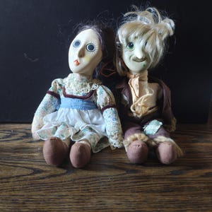 May include: A pair of vintage dolls, one with a green face and blonde hair, the other with a pink face and brown hair. Both dolls are wearing clothing and sitting on a wooden surface.