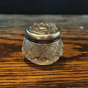 May include: A small, antique glass jar with a silver-coloured lid. The jar has a cut-glass design, and the lid is ornate with a floral pattern. The jar is likely used for storing small items, such as cosmetics or jewellery.