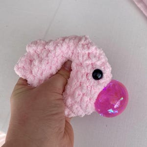May include: A handmade pink plush toy with a black button eye and a large, iridescent pink bubble-shaped nose. The plush is held in a hand against a white background. The toy has a soft, fuzzy texture.