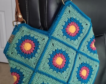 May include: A crocheted tote bag with a teal, blue, pink, and yellow floral pattern. The bag has brown leather handles.
