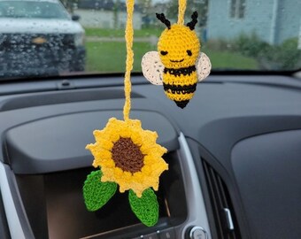 Op de afbeelding: Een gehaakte zonnebloem en bij hangen aan de achteruitkijkspiegel van een auto. De zonnebloem is geel met een bruin hart en groene bladeren. De bij is geel en zwart met witte vleugels.