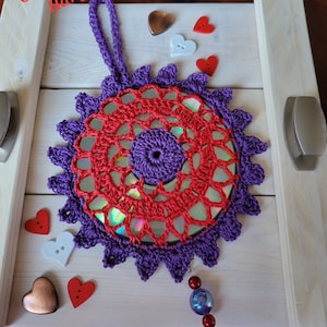 Può includere: Un ornamento appeso a forma di CD in crochet viola e rosso con una corda di perline che pende dal fondo. L'ornamento è appeso su una superficie di legno bianco con cuori rossi e bianchi.