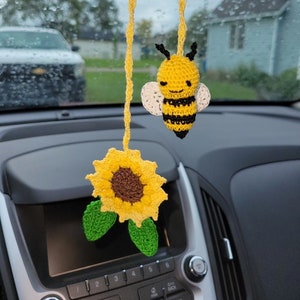 May include: A crocheted sunflower and bee hanging from the rearview mirror of a car. The sunflower is yellow with a brown center and green leaves. The bee is yellow and black with white wings.
