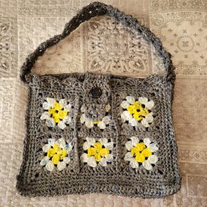May include: A handmade gray crochet tote bag with a daisy pattern. The bag features a button closure and a braided strap. The daisy pattern has yellow and white flower centers. The bag is made of a textured yarn.