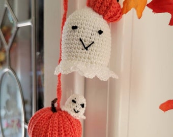 May include: A crochet hanging decoration featuring a white ghost with a black stitched face and a ruffled bottom edge, paired with a small orange pumpkin. A second, smaller white ghost sits atop a larger orange pumpkin, all connected by a red yarn cord.