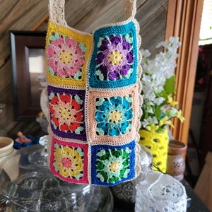 May include: A colorful crochet bag with a cream-colored strap. The bag is made of squares in various colors, including pink, yellow, blue, and purple, each featuring a floral design. The edges are trimmed with a scalloped cream border.