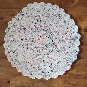 May include: A round, handmade crochet rug with a scalloped edge. The rug is primarily white with flecks of pink, black, and blue throughout. The design spirals from the center outwards. The rug is placed on a wooden floor.