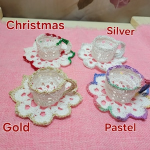 Könnte beinhalten: Vier gehäkelte Teetassen und Untertassen in verschiedenen Farben: Weihnachten (rot und grün), Silber (weiß), Gold (gold) und Pastell (lila, rosa und grün).