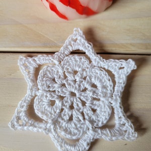 May include: A white, crocheted snowflake ornament with a six-pointed star design. The ornament is placed on a light wood surface, with a red and white striped candle in the background. The snowflake is a handmade item.