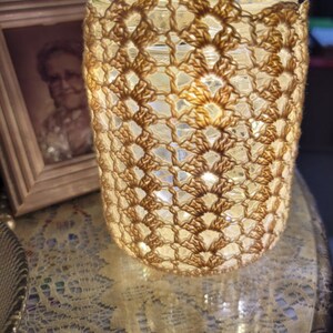 May include: A crocheted jar cover with a handle, made with beige yarn. The cover is over a glass jar with string lights inside, creating a warm glow.
