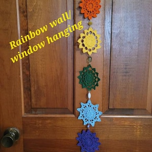 May include: Rainbow wall/window hanging made of crocheted stars in different colors, including red, orange, yellow, green, blue, and purple. The stars are connected with beads and hang from a wooden door.