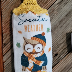 May include: A white kitchen towel with a graphic of an owl wearing a blue hat and a plaid scarf, surrounded by autumn leaves. The text "Sweater Weather" is printed above the owl. A yellow crocheted topper with a brown button hangs from a rod.