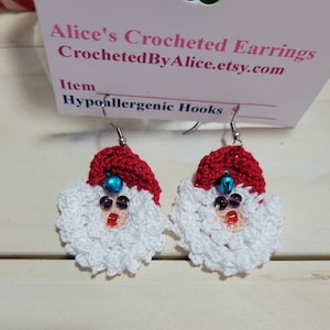 May include: A pair of crocheted Santa Claus earrings. Each earring features a red hat, white beard, and a face with bead accents. The earrings are suspended from silver-colored hooks. The tag reads "Alice's Crocheted Earrings".