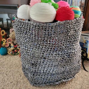 May include: A gray woven basket filled with various colors of yarn. The yarn includes white, pink, green, red, and rainbow-colored skeins. The basket is sitting on a beige carpet.