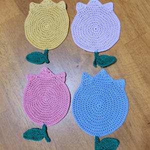 May include: Four handmade crochet tulip-shaped coasters in yellow, lavender, pink, and blue. Each coaster has a green leaf and stem detail. The coasters are arranged on a wooden surface, showcasing their vibrant colours and floral design.
