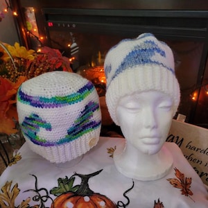 Puede incluir: Dos gorros tejidos, uno blanco con un patrón de olas azul, verde y morado, y otro blanco con un patrón de olas azul. Los gorros cuentan con un borde acanalado y una pequeña etiqueta circular. Un gorro se muestra en un maniquí blanco.