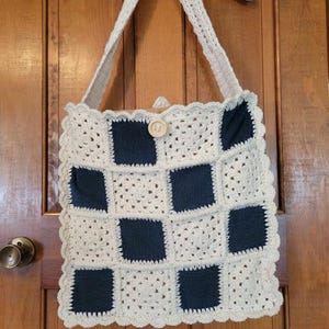 May include: A handmade crochet tote bag with a checkerboard pattern. The bag features alternating cream-colored and navy blue squares, a cream-colored scalloped edge, and a wooden button closure. The bag has a cream-colored strap.