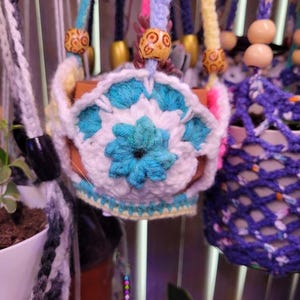 May include: A crochet plant hanger featuring a white and turquoise floral pattern with a central blue flower motif. The hanger includes light blue yarn straps adorned with round wooden beads featuring dark brown circular patterns.