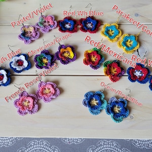 May include: Crocheted flower earrings in various colors, including pastel violet, red, white, blue, grape, French rose, parakeet gold, and ocean blue. The earrings are displayed on a wooden surface.