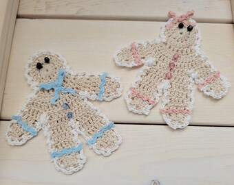 May include: Two crocheted gingerbread man ornaments. One has a blue bow and trim, the other has a pink bow and trim. Both have black bead eyes and button details. The ornaments are beige with white scalloped edges.