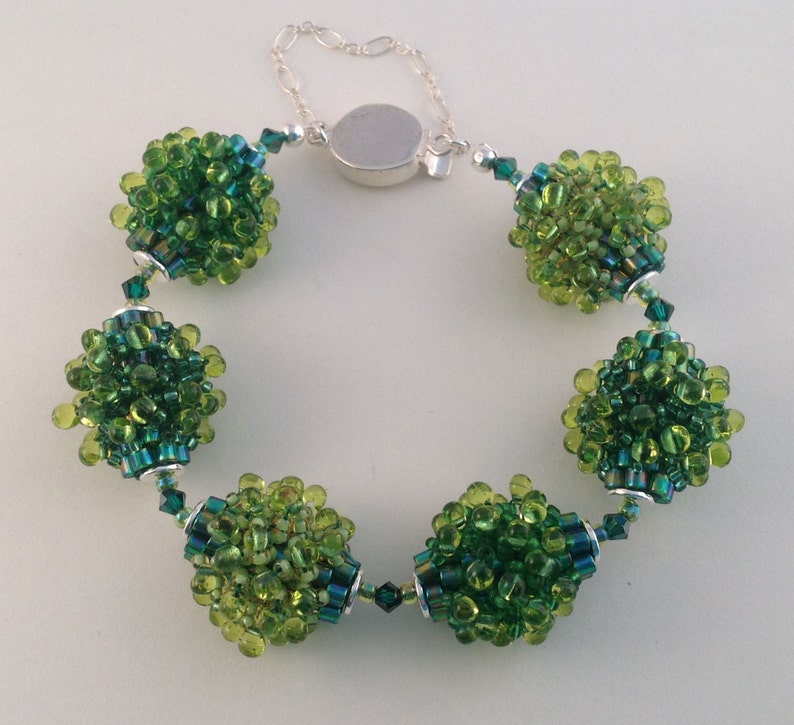 May include: A beaded bracelet with clusters of green beads in different shades, linked by silver-coloured metal accents and a chain clasp. The bracelet has a unique, textured look with a mix of light and dark green beads.