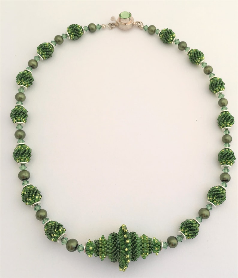 Pu&ograve; includere: Una collana di perline verdi con un grande ciondolo di perline verdi. La collana &egrave; fatta di piccole perline verdi e perline d'argento. Il ciondolo &egrave; fatto da un gruppo di perline verdi.